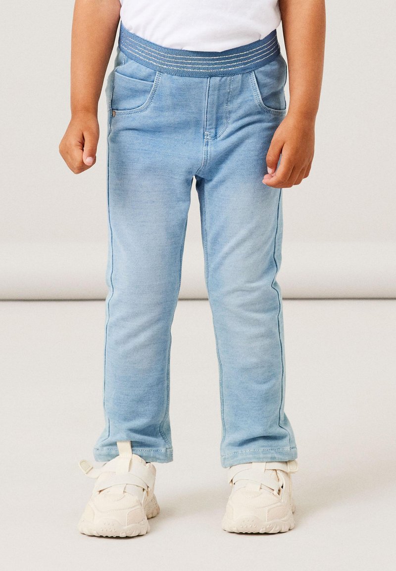 Light blue denim pants with a stretchy waistband, front pockets, and a slim fit. Paired with white sneakers featuring adjustable straps.