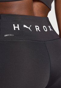 Black athletic leggings with a high waistband, featuring white "H Y R O X" text and Puma logo. Material is smooth with a dry-fit texture.