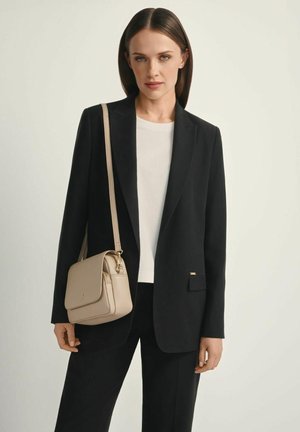 Kobieta ubrana w czarną marynarkę, biały top i czarne spodnie, niosąca beżową torbę na ramię z długim paskiem.