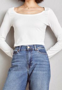 Vit långärmad topp med fyrkantig halsringning, matchad med högmidjade blå jeans med knappstängning och subtila blekningar.