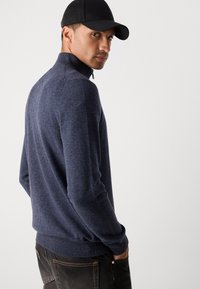 Navy knit sweater with high collar and half-zip closure, featuring ribbed cuffs and hem, worn with dark jeans.