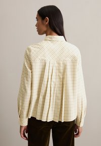 Yellow checkered blouse with a relaxed fit, featuring a pleated back design and long sleeves, paired with brown corduroy pants.