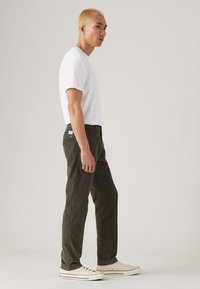 Young man with short blonde hair wearing a white t-shirt, dark green pants, and white sneakers standing sideways on a plain background.