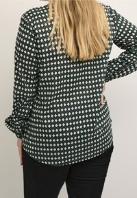 Langsleeve shirt in donkergroen met een ruitpatroon in wit en grijs. Kenmerkt zich door een gebogen zoom en twee zijzakken.