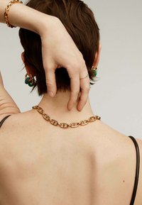 Collar de cadena de oro con eslabones alargados, llevado en la parte posterior del cuello, combinado con pendientes verdes gruesos y una camiseta de tirantes negra.