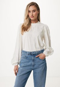 Blusa bianca a maniche lunghe con ricami floreali e maniche a sbuffo. Abbinata a jeans blu a vita alta, mostrata in una posa casual in piedi.