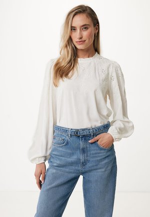 White, long-sleeve blouse with floral embroidery and puff sleeves. Paired with high-waisted blue jeans, featured in a casual standing pose.