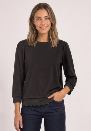 Femme aux cheveux mi-longs sourit, portant un haut noir à manches longues avec un ourlet en dentelle festonnée et un jean bleu foncé, main dans la poche.