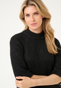 Schwarzer Strickpullover mit hohem Kragen, halblangen Ärmeln und einer strukturierten Oberfläche. Der Stoff scheint weich und gemütlich zu sein.