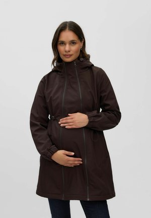 Femme enceinte portant une veste de maternité marron foncé avec capuche, tenant doucement son ventre devant un fond clair uni.