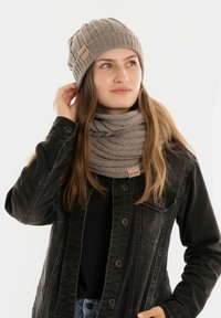 Gebreide beanie en sjaal set in zachte beige tinten; ribbelstructuur met merkelabels. Zwarte denimjack maakt de look compleet.