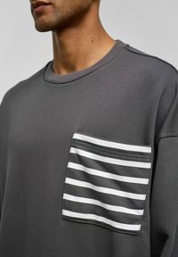 Grijze crewneck sweatshirt met een gestreepte zak in wit en grijs. Gemaakt van lichtgewicht stof, met een losse pasvorm en zichtbare stiksels.