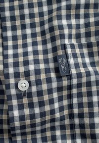 Camisa a cuadros en azul marino y beige con un botón blanco, bolsillo cosido y un pequeño parche con un diseño de astas.