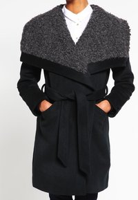 Cappotto di lana nero con colletto grigio peloso, design avvolgente, maniche lunghe, vita con cintura, tasche laterali e una consistenza morbida.