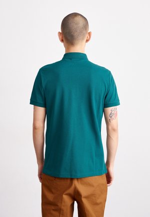 Mann mit rasiertem Kopf, der ein türkisfarbenes kurzärmeliges Poloshirt und braune Hosen trägt, von hinten gesehen, mit einem Tattoo am linken Oberarm.