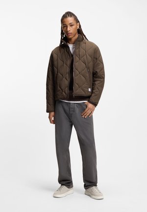 Ruskea tikattu takki, jossa korkea kaulus ja etuvetoketju, yhdistetty harmaisiin housuihin ja vaaleisiin lenkkareihin. Tasainen pinta ja hillityt design-yksityiskohdat.