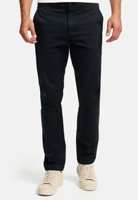 Smaragdgrüne Chinos aus einer Baumwollmischung, mit seitlichen und hinteren Taschen, einem Reißverschluss und einem slim fit Design. Getragen mit beigen Sneakers.