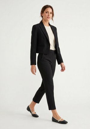 Kvinde iført sort cropped blazer, sorte slim bukser, cremefarvet top og sorte flade sko med cremefarvede stropper mod en ensfarvet hvid baggrund.
