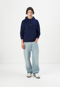 Navy blue hoodie with drawstrings and kangaroo pocket, paired with light blue, loose-fitting jeans, and white sneakers with black accents.