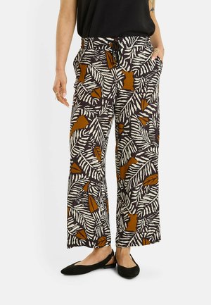 Mujer que lleva pantalones de pierna ancha con patrones abstractos de hojas en negro, blanco y marrón, combinados con bailarinas negras y una camiseta negra.