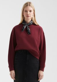 Felpa bordeaux con collo a giro e spalle cadute, abbinata a un bandana nero. L'abbigliamento presenta una vestibilità comoda e dettagli minimi.