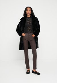 Kvinde iført en sort faux fur frakke over en sort blonde top, brune bukser og sorte strukturerede slip-on sko, stående foran en ensfarvet hvid baggrund.