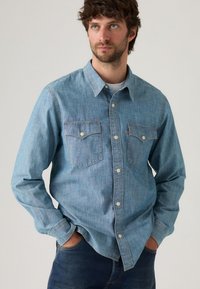 Light blue denim shirt with long sleeves, button-down collar, and two front pockets. Features contrasting white buttons and a soft texture.
