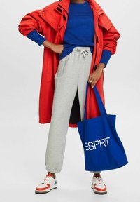 Rote Kapuzenjacke, blauer strukturierter Pullover, graue Jogginghose, weiße Sneaker mit roten Akzenten und eine blaue Tragetasche mit dem "ESPRIT"-Logo.