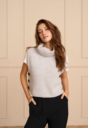 Une femme pose les mains dans les poches. Elle porte un haut blanc en maille, sans manches et à col bénitier, par-dessus une chemise blanche et un pantalon noir.