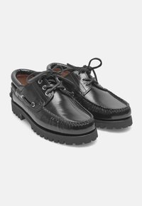 Schwarze Lederschuhe mit Mokassin-Design, ausgestattet mit geflochtenen Nähten, runden Schnürsenkeln und einer strukturierten Gummisohle. Akzente umfassen metallische Ösen.