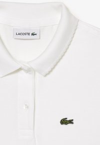 Biała koszulka polo z teksturowanej tkaniny, z trzema guzikami na plisie oraz haftowanym zielonym logo krokodyla po lewej stronie klatki piersiowej.