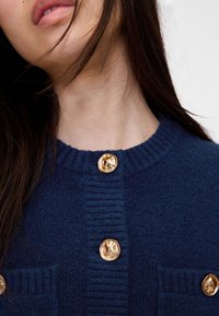 Tamsiai mėlynas megztinis iš mezgimo su ribbais kaklu ir tekstūruota medžiaga, puoštas auksinių atspalvių sagomis ir dviem priekinių kišenių.