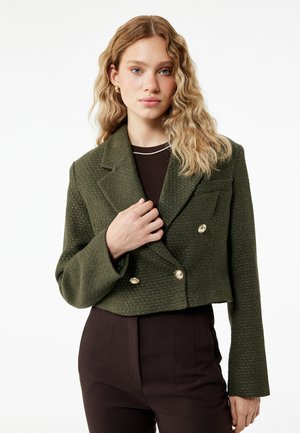 Junge Frau mit welligem blondem Haar, die einen kurzen olivgrünen strukturierten Blazer mit goldenen Knöpfen über einem dunklen Oberteil und Hose trägt.