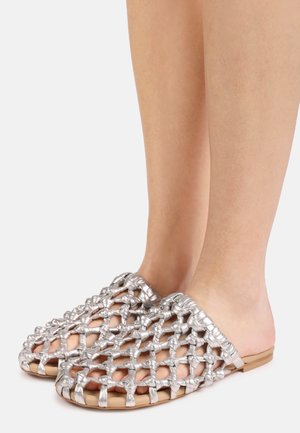 Füße, die Slip-On-Schuhe mit breiten, offen gewebten silbernen Riemen und braunen flachen Sohlen auf einem einfarbigen hellen Hintergrund tragen.