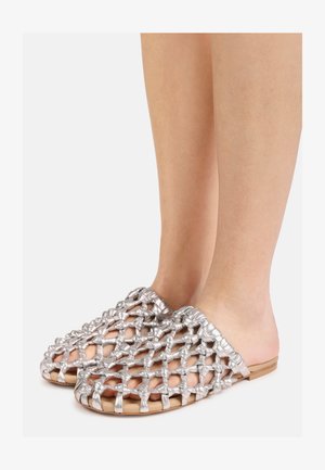 Pies con zapatos sin cordones con tiras anchas plateadas de tejido abierto y suelas planas marrones sobre un fondo claro liso.