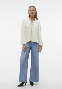 Blouse crème clair avec un devant froncé, manches longues, associée à un jean bleu clair à jambes larges et des escarpins noirs à bout pointu. Design simple et texturé.