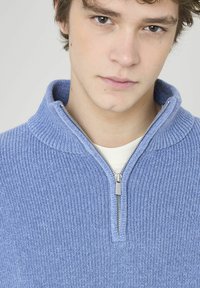 Pullover en tricot côtelé bleu clair avec un col zippé haut. Présente une fermeture éclair argentée, porté sur une chemise blanche. Texture douce. Vue en gros plan.