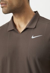 Gros plan d'un homme portant un polo marron texturé Nike avec un logo Nike blanc sur la poitrine.