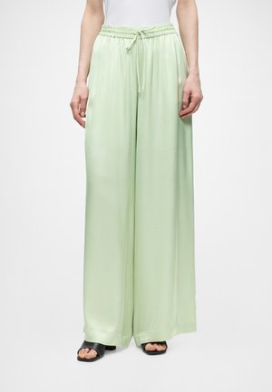 Γυναίκα που φοράει ανοιχτό πράσινο παντελόνι με φαρδιά πόδια, ελαστική μέση και κορδόνι, συνδυασμένο με μαύρα σανδάλια ανοιχτής μύτης, στέκεται σε λευκό φόντο.