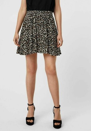 Vrouw met een korte rok met luipaardprint en zwarte hoge hakken met enkelbandje, staand tegen een effen achtergrond.