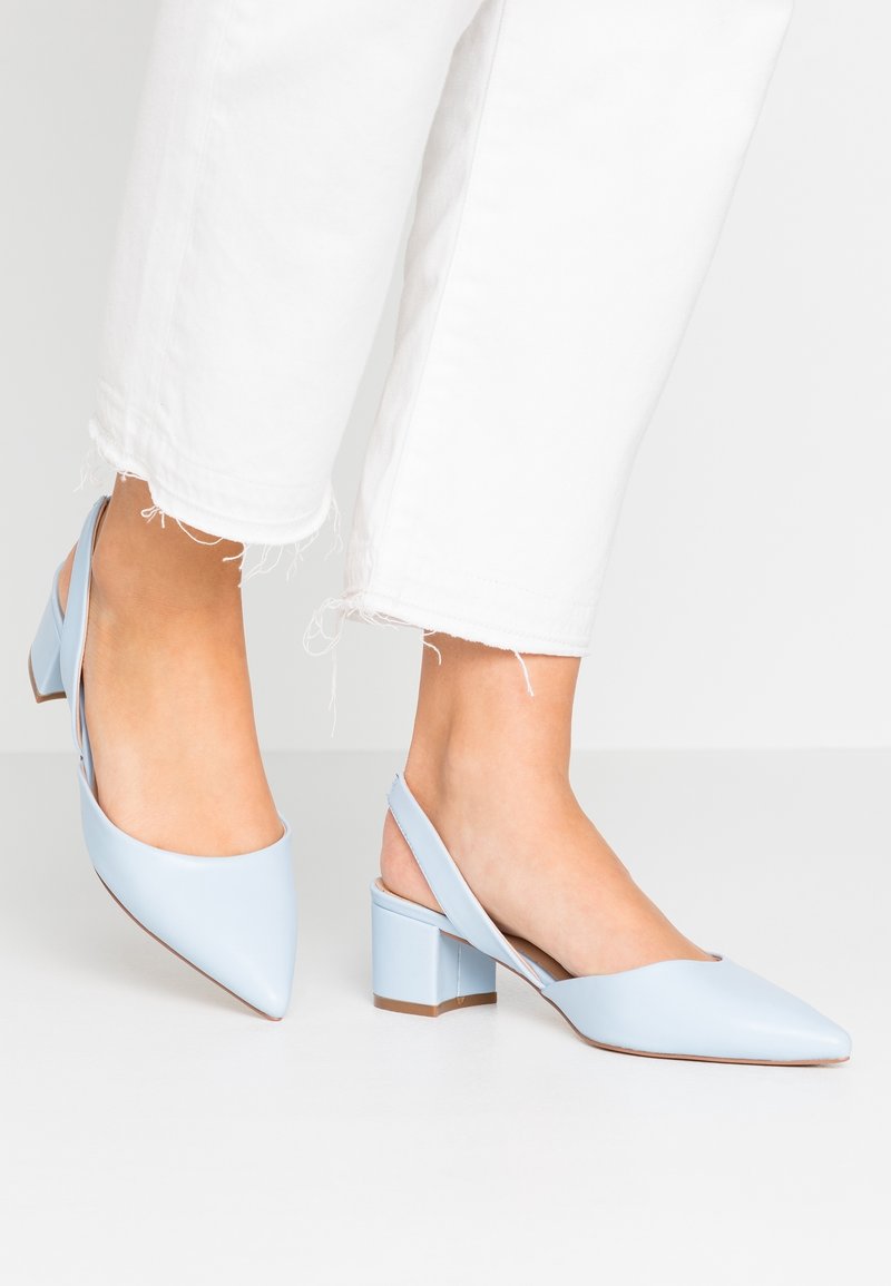 Femme portant des chaussures à talon bloc bleu clair avec bride arrière, bouts pointus, et pantalon blanc court à ourlet effiloché.