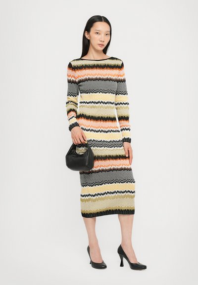 Abito in maglia con motivo zigzag multicolore in arancione, nero, verde e giallo. Maniche lunghe e silhouette aderente, indossato con tacchi neri e borsa.
