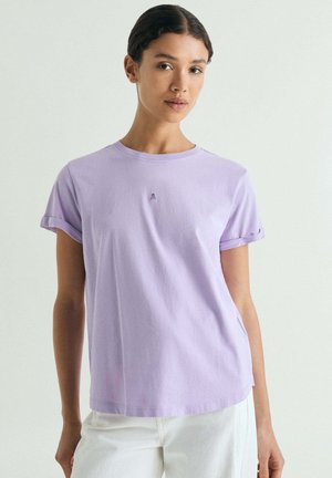 Mujer con una camiseta de manga remangada de color púrpura claro y pantalones blancos, de pie frente a un fondo claro y liso.