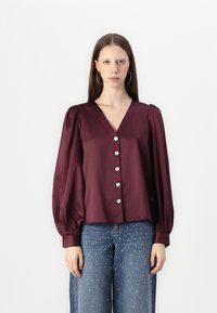 Burgundy satin blouse with a V-neck, puffed sleeves, and five floral buttons down the front. Paired with light blue jeans featuring embellishments.