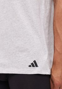 Chemise grise à manches courtes en tissu doux, avec un accent de logo noir en bas à gauche de l'ourlet. Texture lisse et coupe décontractée.