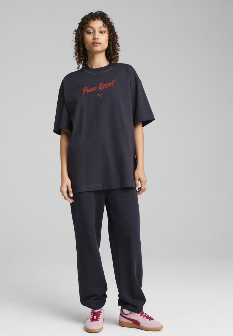 Jūrų laivyno per didelis marškinėliai su raudonu "Puma Resort" logotipu, derinami su tamsiomis kelnėmis ir rožinėmis sportinėmis bateliais su raudonomis detalėmis ir šviesiai rudiems padu.
