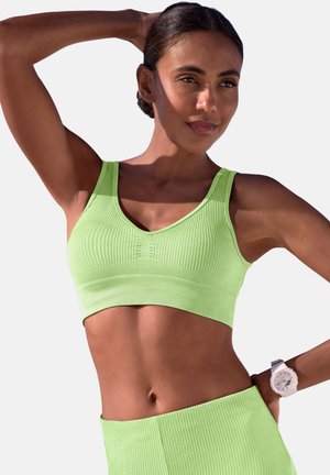 Frau mit dunklen Haaren in einem limettengrünen, gerippten Sport-BH und Leggings, die mit einem erhobenen Arm posiert und eine weiße Armbanduhr trägt.