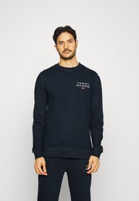 Sweatshirt bleu marine à manches longues, col rond et logo sur la poitrine, fabriqué dans un tissu doux. Pantalon assorti visible en dessous.