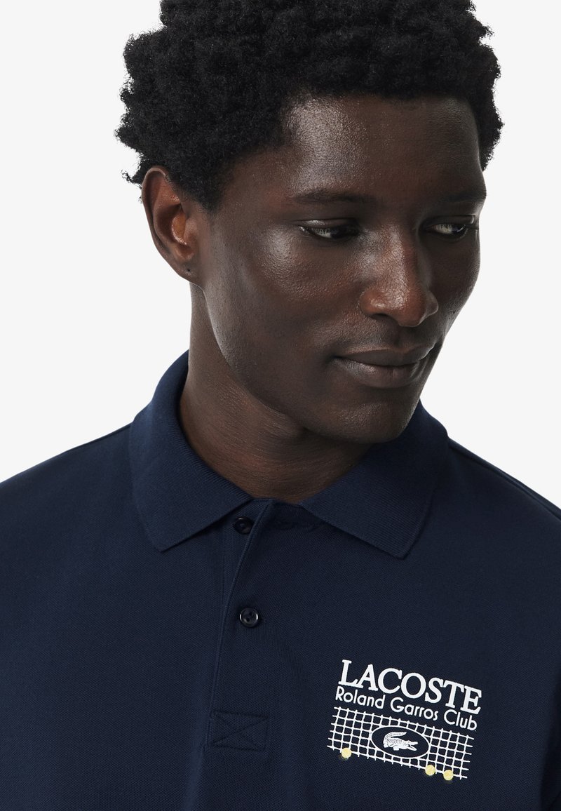 Homme aux cheveux bouclés portant un polo bleu marine Lacoste Roland Garros Club, regardant vers le bas, tourné vers la droite.