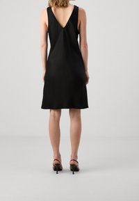 Schwarzes ärmelloses Kleid mit V-Ausschnitt, glatter Textur, knielangem Schnitt und leichter A-Linie. Getragen mit Riemchenheels.